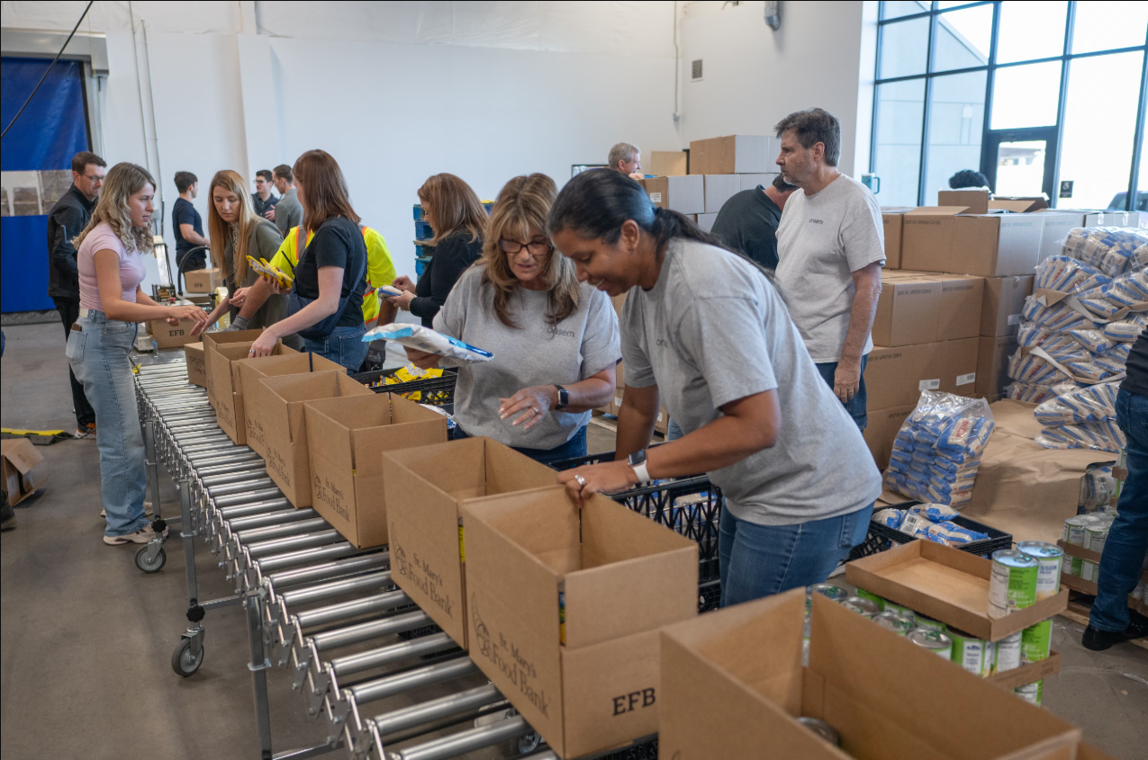 A group of volunteers work together in a warehouse, packing food and supplies into boxes on a conveyor system for community assistance and donation efforts.